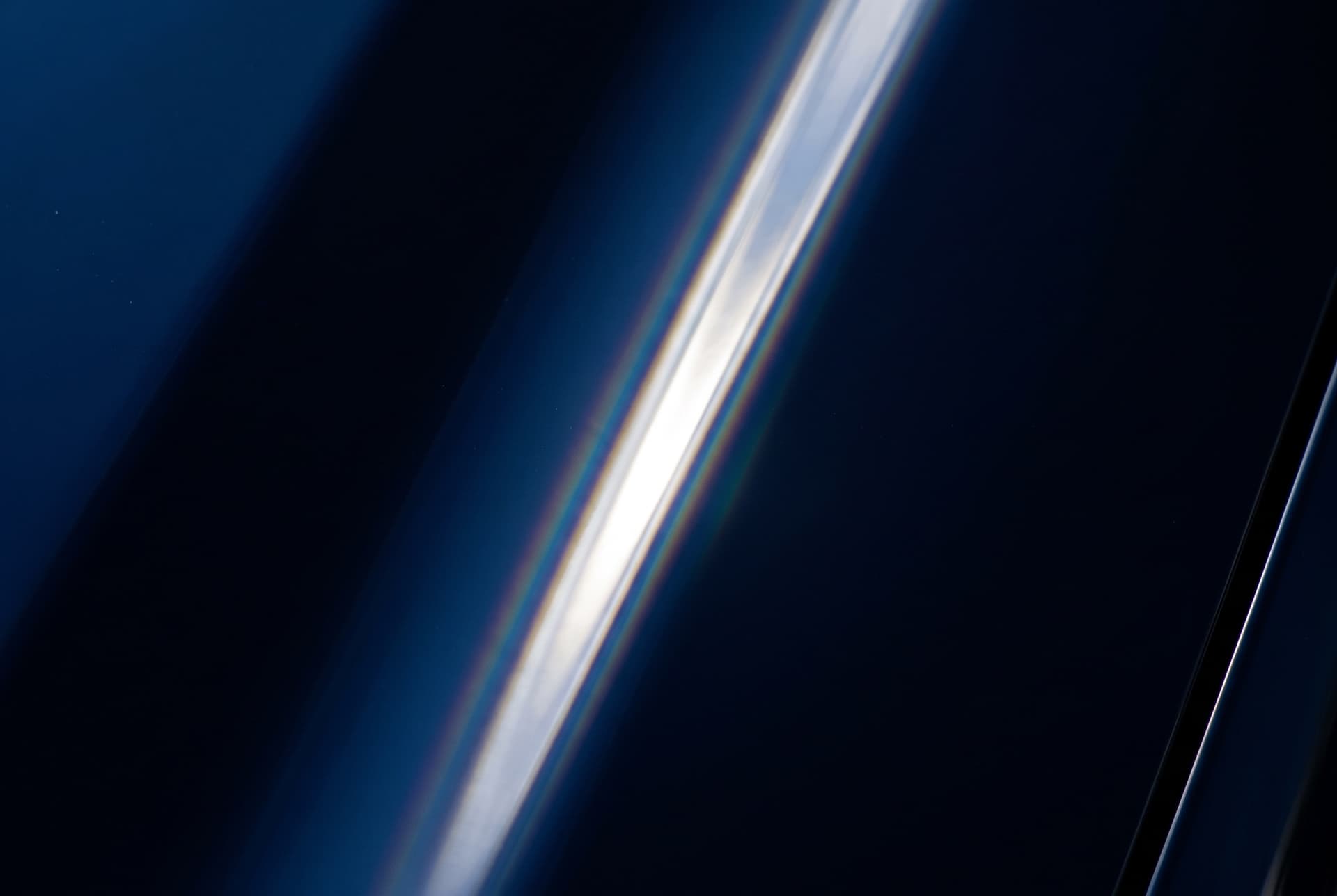 A hard line of light travelling across flawless dark blue car paint, the highlight edged with a faint warm and cool fringe